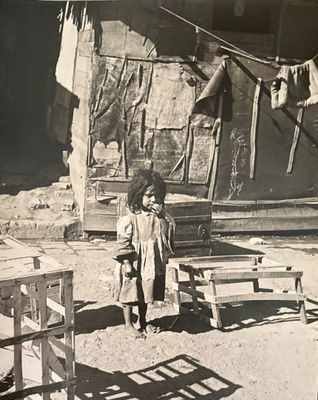 a black and white photo of a little girl standing in front of a shack