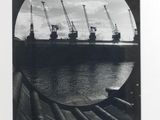 black and white photograph of a ship in the water with cranes in the background