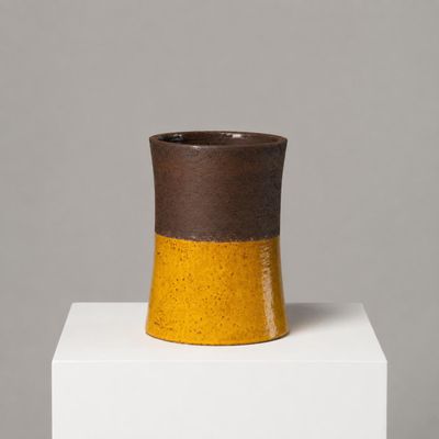 vase en céramique marron et jaune posé sur un socle blanc, sur fond gris
