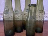 six bouteilles en verre vert vides posées sur une table en bois près d'un mur blanc
