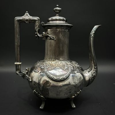 théière plaquée argent, ornée de motifs élaborés, posée sur un socle, sur fond noir