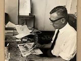 photographie en noir et blanc d'un homme assis à un bureau, entouré de papiers