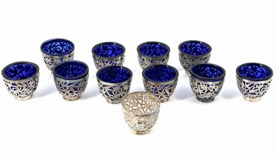 six blue glass cups and a silver vase on a white surface