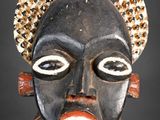 une image représentant un masque en bois au visage noir et coiffé d'une parure dorée