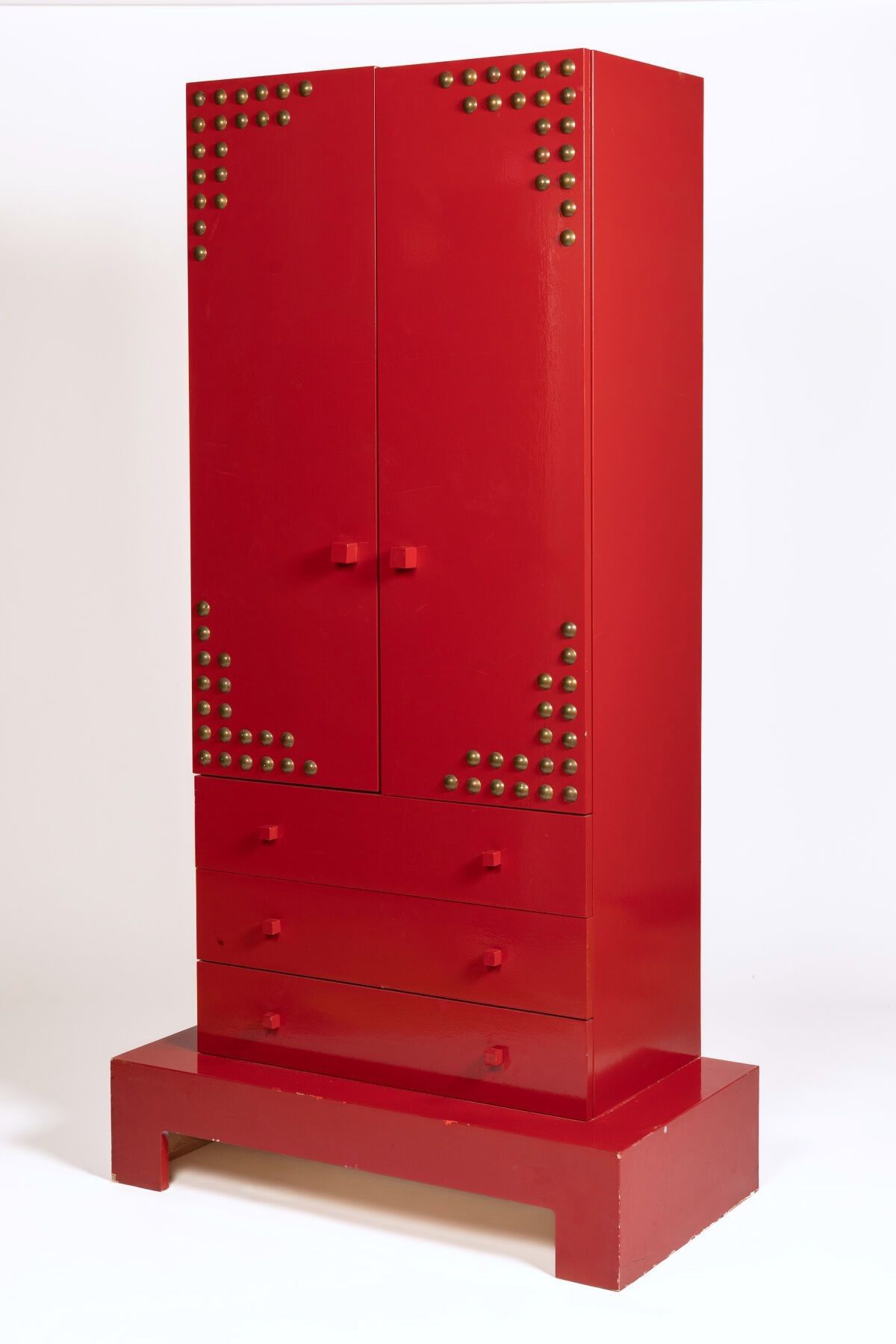 Armoire en bois laqué rouge, ouvrant à deux vantaux cloutés découvrant des étagères en partie haute, et par trois tir...
