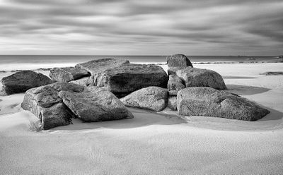 photographie en noir et blanc représentant des rochers sur une plage, sous un ciel nuageux