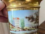 a hand holding a gold vase with a painting on it