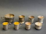 six tasses en argent munies d'anses sont alignées sur une surface grise