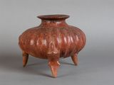 un petit vase rouge posé sur un socle, sur une surface grise