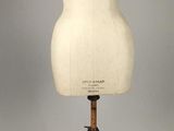 une image représentant un mannequin posé sur un socle en bois, sur fond gris