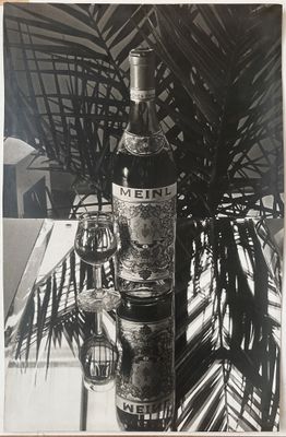 photographie en noir et blanc d'une bouteille de vin posée sur une table à côté d'un palmier