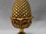 une pomme de pin dorée posée sur un socle, sur fond blanc