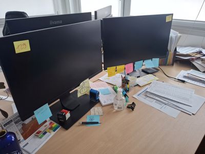 two computer monitors on the desk with papers on the desk