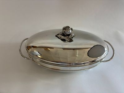 une photo représentant un plat en argent muni d'un couvercle, posé sur une table blanche