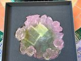emballage d'une broche en forme de fleur rose, verte et jaune, dans une boîte noire posée sur une table en mosaïque