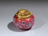un vase rouge avec un couvercle doré posé sur une surface blanche