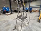 une photo représentant une échelle métallique dans un entrepôt, avec des cartons et des palettes en arrière-plan