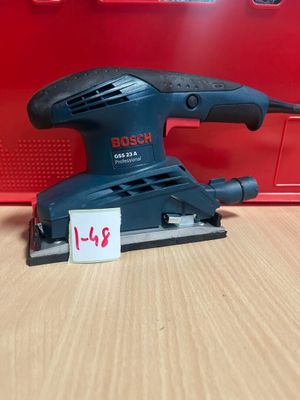 une ponceuse orbitale Bosch posée sur une table en bois, à côté d'une boîte rouge