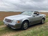 Une Mercedes Classe SL argentée, version cabriolet, garée dans un champ, avec un ciel nuageux en arrière-plan