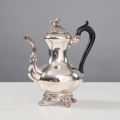 une théière ancienne en argent, avec une anse noire, posée sur une table blanche
