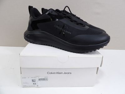 Des baskets noires avec un logo sur la semelle sont présentées dans une boîte blanche.