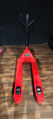 un chariot élévateur rouge posé sur un plancher en bois, à côté d'une paire de pieds