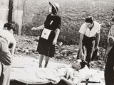 photo en noir et blanc d'un groupe de personnes debout autour d'un homme allongé sur un lit