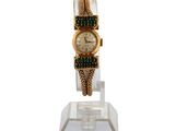 une montre en or ornée de pierres vertes, posée sur un socle transparent