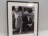 photographie en noir et blanc d'une femme appuyée contre une voiture, à côté d'un homme appuyé contre l'arrière de celle-ci
