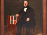portrait d'un homme en costume noir et nœud papillon, assis sur une chaise