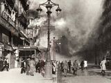 black and white photograph of people walking down a street with a fire in the background
