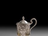 une théière ancienne en argent, avec un couvercle et une anse, posée sur un support gris