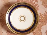 une assiette bleue et dorée, bordée d'or, posée sur un papier peint à motifs floraux