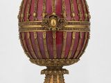 un œuf richement décoré de rouge et d'or, posé sur un socle, sur fond blanc