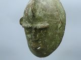 un masque antique en bronze posé sur un socle, sur fond gris
