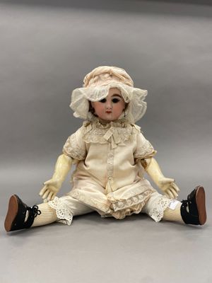 une poupée en porcelaine ancienne, assise par terre, vêtue d'une robe blanche et d'un bonnet