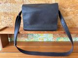 black leather messenger bag sitting on a wooden bench in front of a brick wall