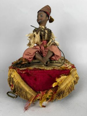 une vieille poupée assise sur un coussin rouge, une pipe à la bouche