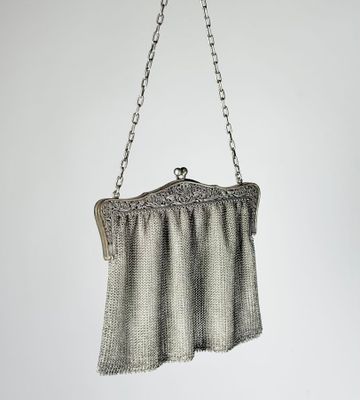 an antique silver mesh purse hanging from a chain on a white wall