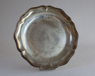 assiette en argent plaqué à bord festonné, posée sur un socle, sur fond blanc