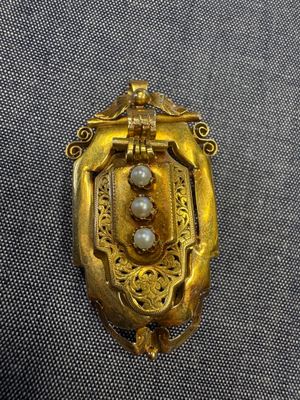 une photo d'une broche en or et perles posée sur un tissu gris