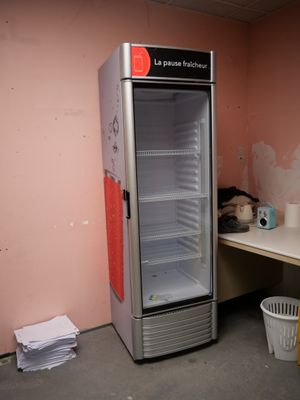 un réfrigérateur vide dans un coin de la pièce, à côté d'une poubelle