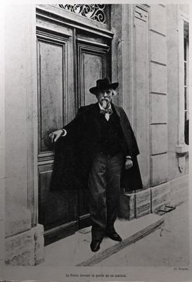 photographie en noir et blanc d'un homme portant un haut-de-forme et un manteau, adossé à une porte