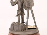 une statue représentant un homme muni d'un appareil photo, posée sur un socle en bois