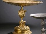two silver and gold plate on a pedestal next to a silver bowl
