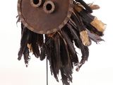 un masque marron orné de plumes, posé sur un socle, sur fond blanc