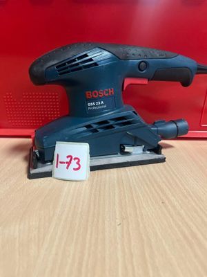 une ponceuse orbitale Bosch posée sur une table, à côté d'une boîte rouge