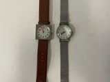 two watches on a white surface one has a brown strap and the other has a silver watch with a silver face