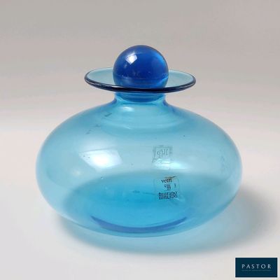 un vase en verre bleu avec un couvercle bleu posé sur une surface blanche