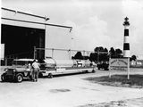 photo en noir et blanc de deux hommes debout à côté d'un camion, avec un phare en arrière-plan